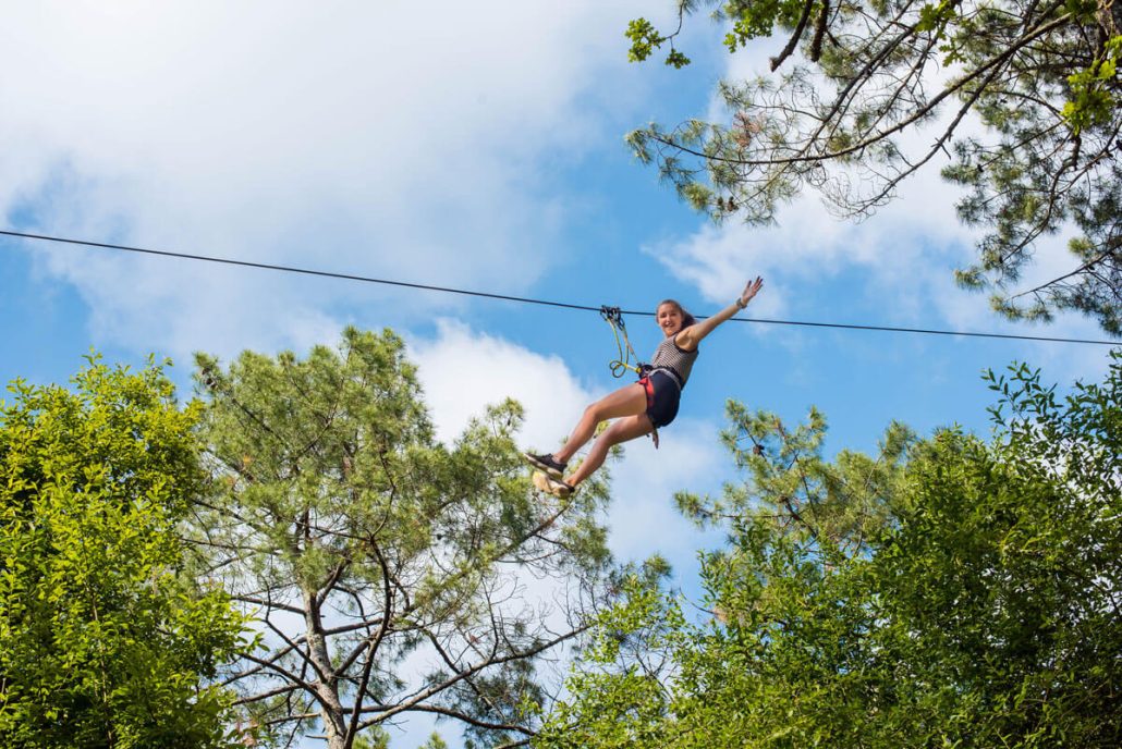 Jeune femme souriante s'élançant sur une tyrolienne géante au-dessus de la forêt de pins au parc d'aventure de La Double Aventure en Charente-Maritime.